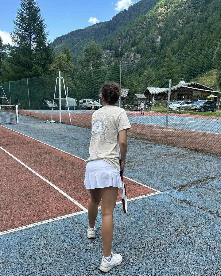 En kvinde på en tennisbane i bjergene, set bagfra i en sandfarvet t-shirt med Metatrons terning sol.